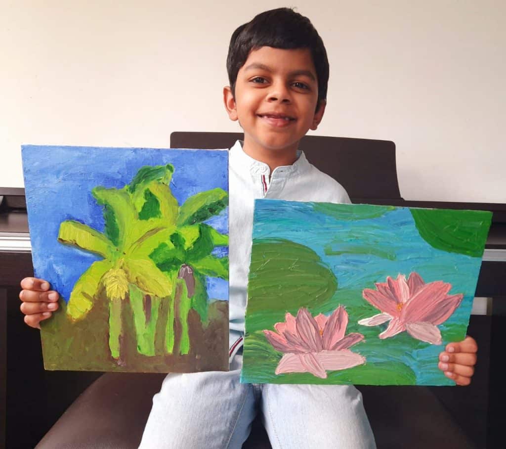 A young boy, Shiven Sidharth, smiles whilst sitting and holding two colourful paintings—one of green banana trees under a blue sky, and the other of pink lotus flowers floating on a pond with green lily pads.