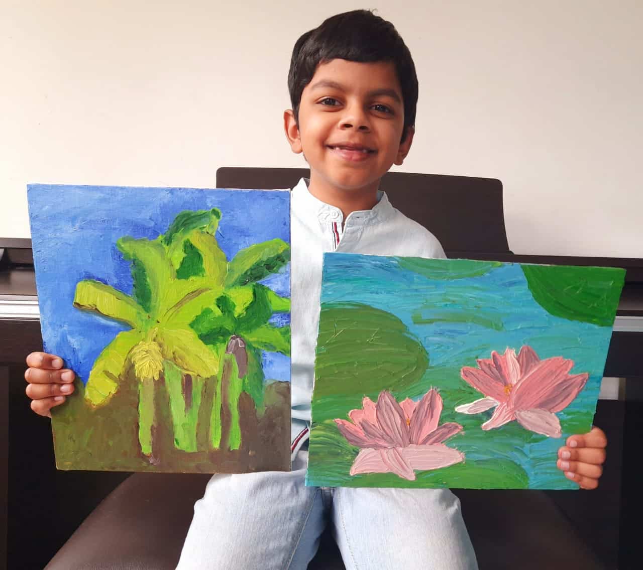 A young boy, Shiven Sidharth, smiles whilst sitting and holding two colourful paintings—one of green banana trees under a blue sky, and the other of pink lotus flowers floating on a pond with green lily pads.