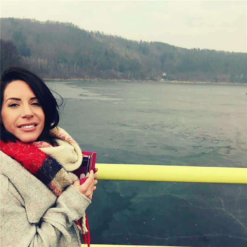 Sophia Barbayannes, in a grey coat and colourful scarf, stands by a yellow railing, smiling at the camera. Behind her is a large, frozen lake with forested hills in the background under a cloudy sky.