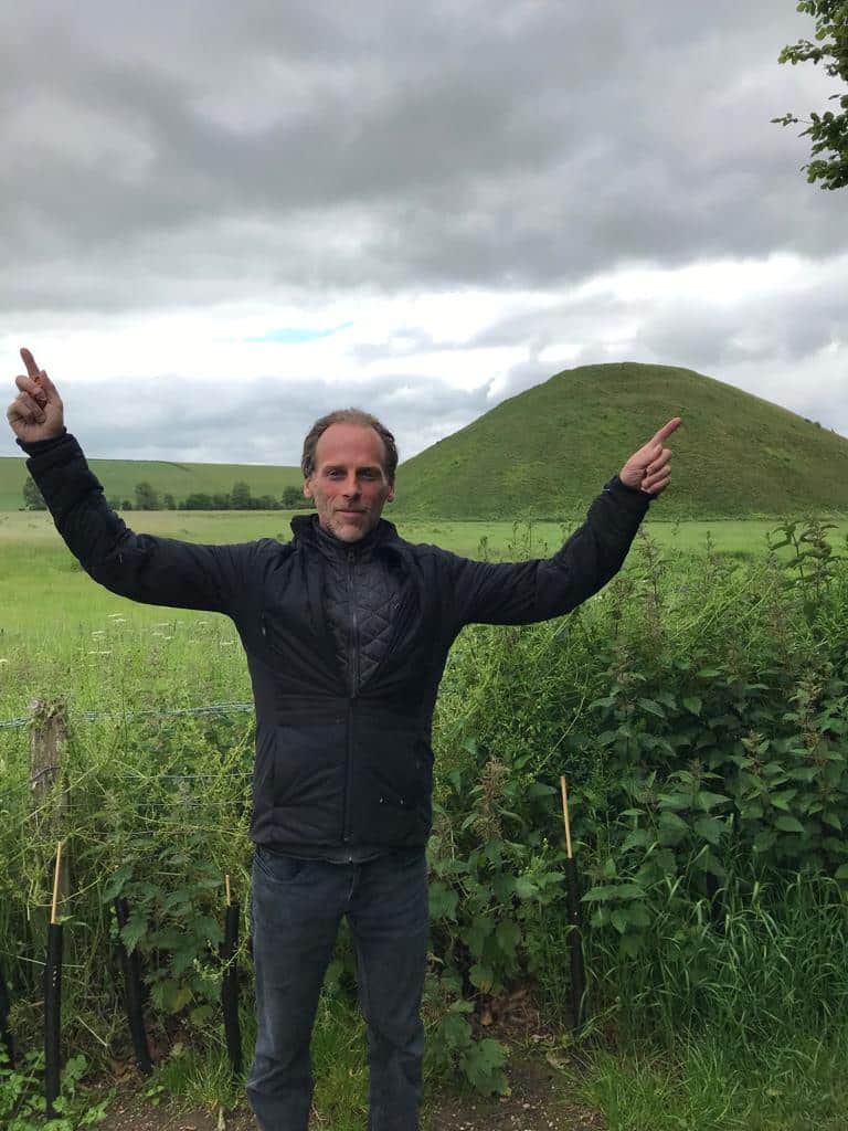 Jim, dressed in a dark jacket, stands outside in a grassy field, raising both arms and pointing upwards, with a large grassy mound and cloudy sky in the background.