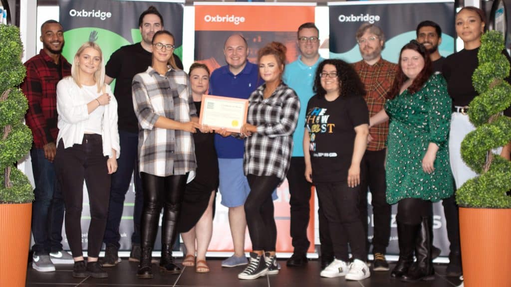 A group of thirteen people stand indoors, posing and smiling. Two women in the front centre hold a certificate. Behind them, Oxbridge banners are displayed, with two tall potted plants flanking the group.