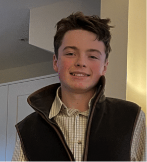 Archie Utting, a teenage boy with short brown hair, smiles at the camera. He is dressed in a checked shirt and dark waistcoat, standing in a softly lit indoor setting with light-coloured walls and cupboards in the background.