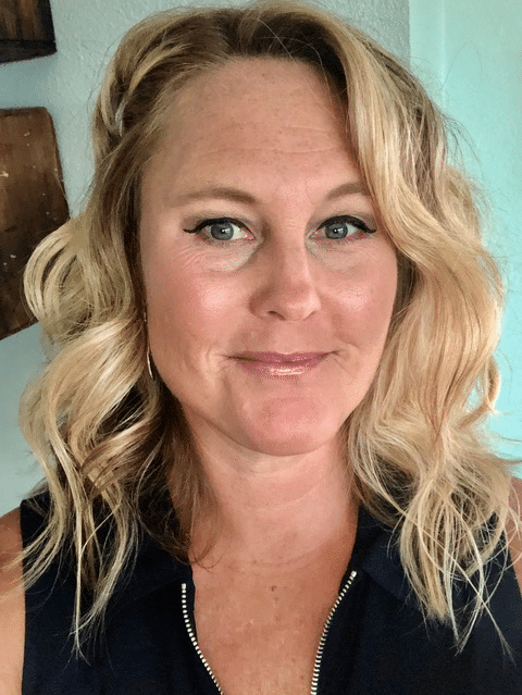 A woman with wavy blonde hair and light eyes smiles slightly at the camera. She is wearing a dark top and subtle make-up, posed indoors against a neutral background that reflects modern interior design aesthetics.
