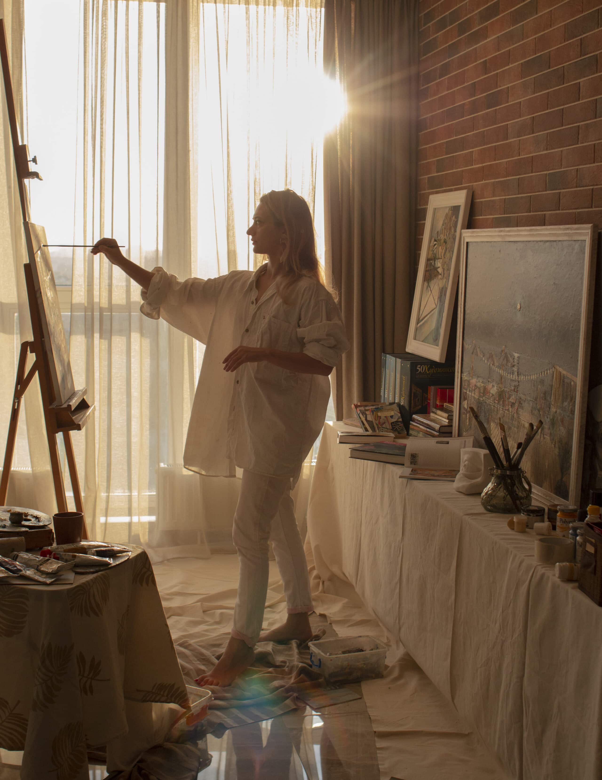 Mariya, in a loose white shirt and trousers, paints on a canvas at an easel in a sunlit room. Art supplies and paintings are scattered around her, with sunlight streaming through the sheer curtains.