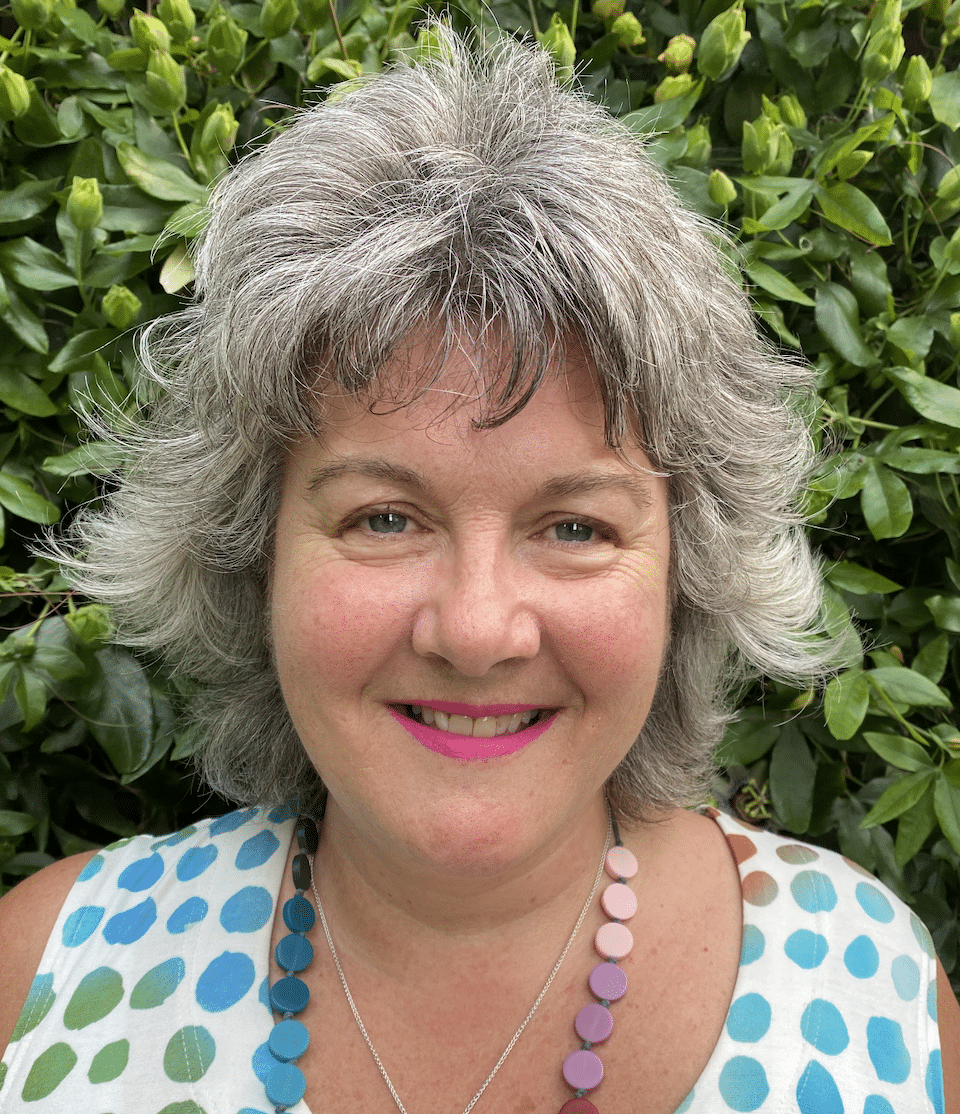 Elaine Stavert, a woman with short, wavy grey hair and pink lipstick, smiles in front of a green leafy background. She wears a sleeveless, polka-dotted top and a colourful beaded necklace.