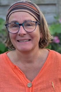 Frances Pitman, a woman with short hair wearing glasses, a striped headband, and an orange shirt, stands outdoors smiling. Green plants and pink flowers create a vibrant backdrop.