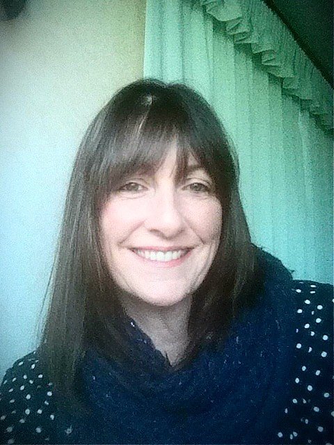 Niki Welch, a woman with straight brown hair and a fringe, smiles at the camera. She is wearing a dark blue scarf and a black top with white polka dots. A light green curtain is in the background.