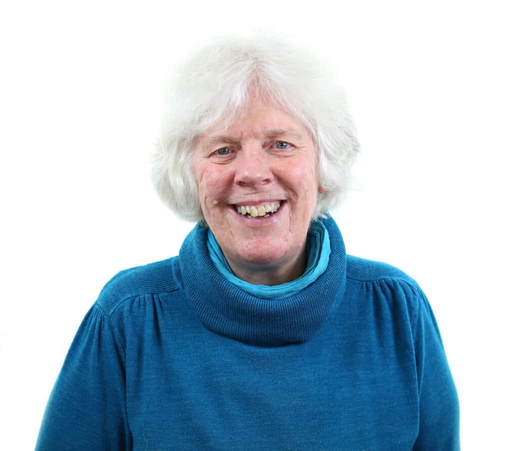 Elaine Watson, an older woman with short white hair, smiles warmly while wearing a teal polo-neck jumper in front of a plain white background.