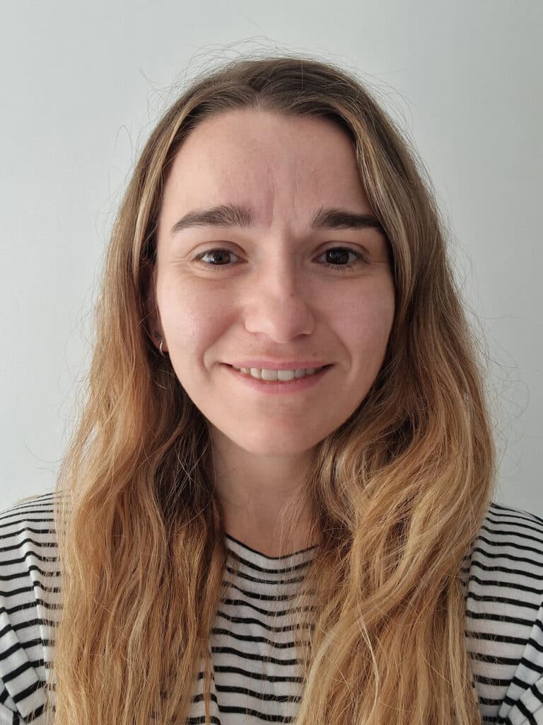 Faye, a young woman with long, wavy blonde hair, is smiling softly. She is wearing a black and white striped jumper and stands in front of a plain white background.