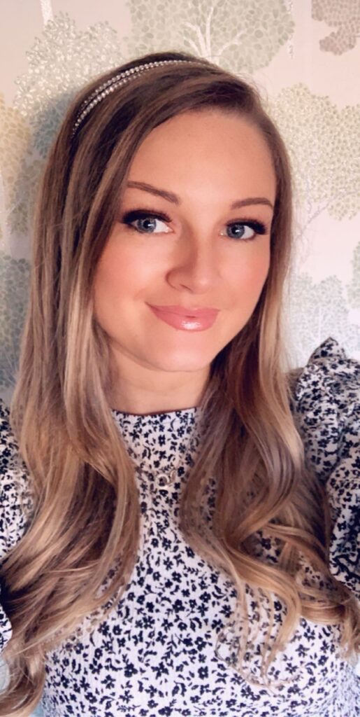 Rebecca Osborne, with long, wavy blonde hair, wears a patterned top and a pearl hairband. She smiles and poses in front of wallpaper featuring light green and white leafy patterns.