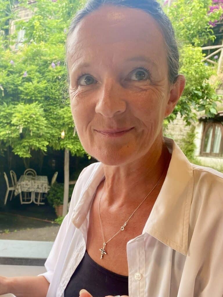 Lisa, a woman with fair skin and short brown hair, wearing a white blouse and a cross necklace, smiles gently whilst sitting outdoors; green foliage and white chairs are visible in the background.