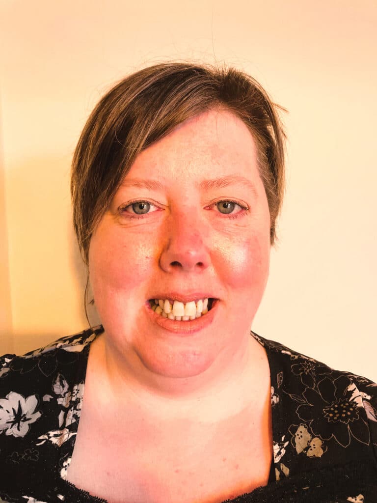 Melanie Lancley, a woman with short brown hair, smiles at the camera. She is wearing a black top with a floral pattern and stands against a light-coloured background.