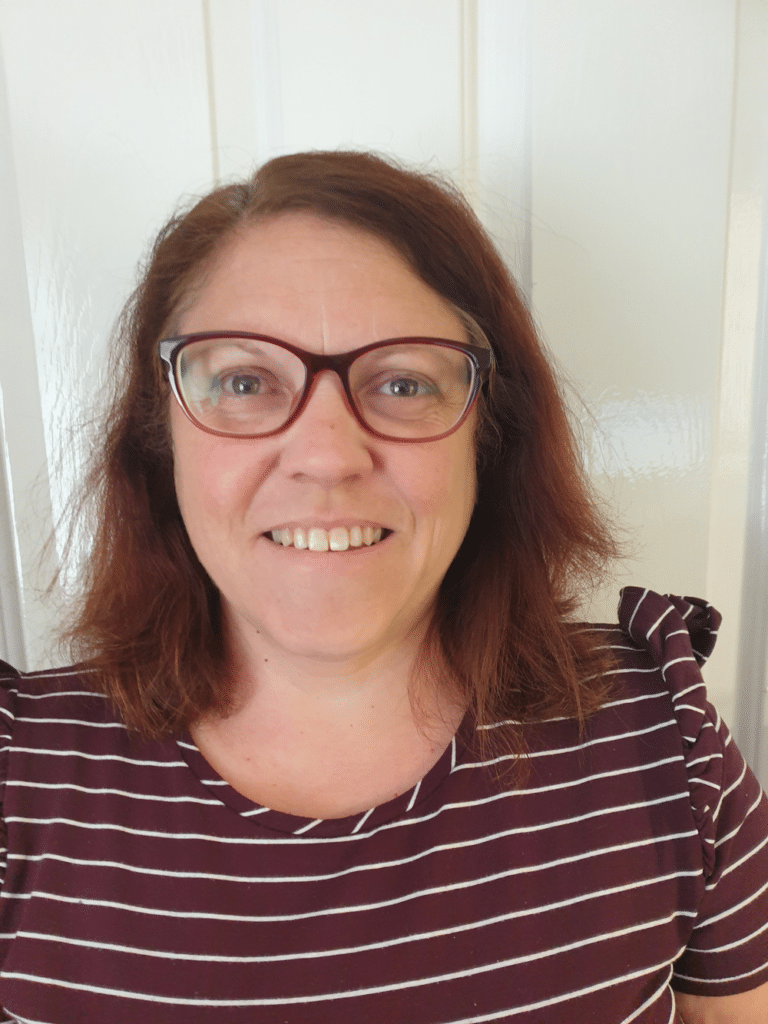 Candice Green, a woman with shoulder-length brown hair and glasses, smiles at the camera. She is wearing a maroon and white striped top, standing in front of a white door.