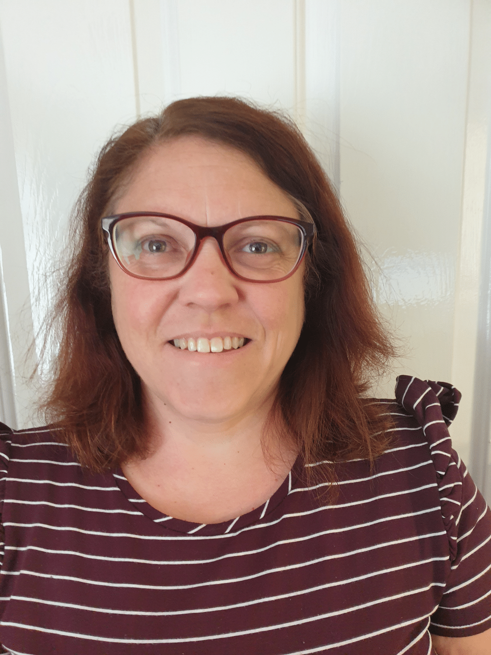 Candice Green, a woman with shoulder-length brown hair and glasses, smiles at the camera. She is wearing a maroon and white striped top, standing in front of a white door.