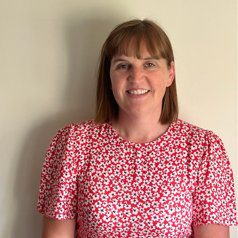 Debbie, a woman with straight brown hair and a fringe, is smiling at the camera. She is wearing a red dress with a white floral pattern and stands in front of a plain, light-coloured wall.