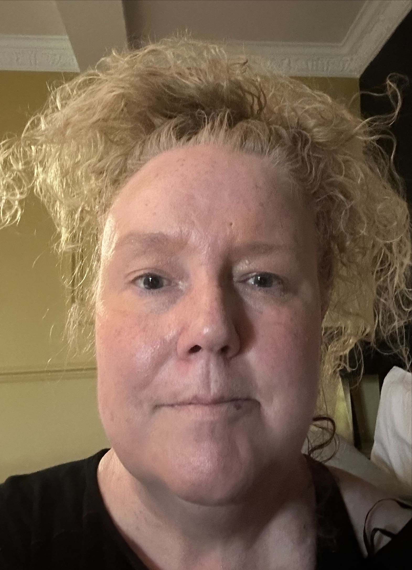Teresa, with fair skin and curly blonde hair, looks directly at the camera in a softly lit indoor setting. Her tousled hair frames her face as she wears a black top against yellow and dark-coloured walls in the background.