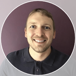 Smiling man with short light brown hair and blue eyes, wearing a dark polo shirt, stands in front of a purple wall, framed by a white circular border—introducing Tim Seed.