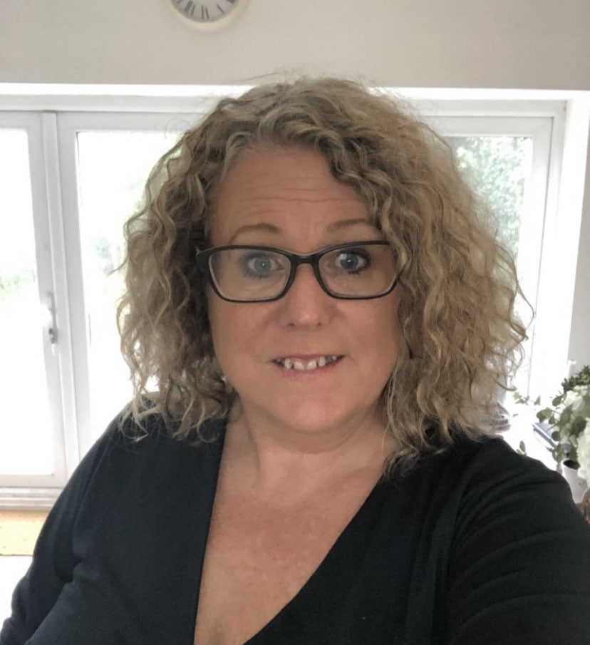 Amanda Trimble, a woman with curly blonde hair, wearing glasses and a black top, smiles at the camera indoors. Large windows and a clock are visible in the background.
