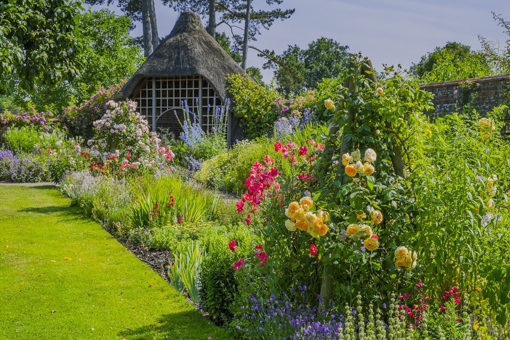 A lush garden designed by Graeme Lisle features colourful flowers and green plants surrounding a thatched-roofed gazebo. A well-maintained lawn leads to vibrant flower beds, with trees and a brick wall in the background under a clear sky.