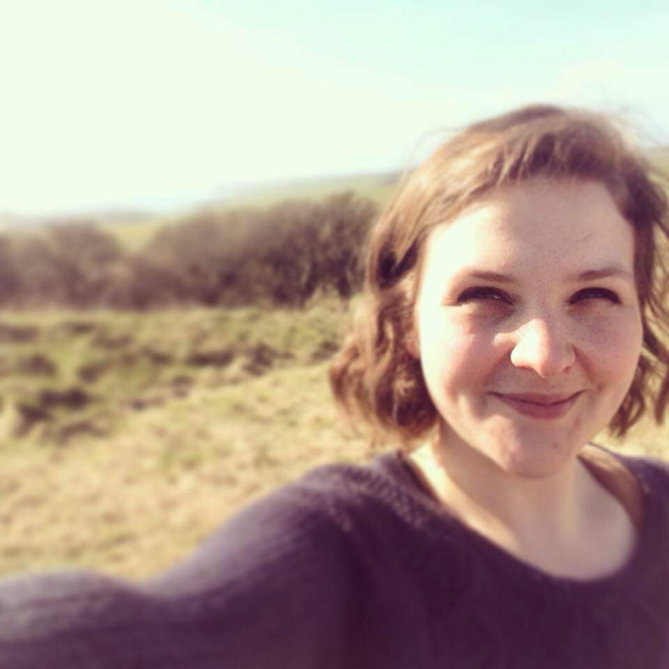 Kat Morgan, a young woman with short, wavy brown hair, smiles while taking a selfie outdoors on a sunny day, with grassy fields and bushes softly blurred in the background.