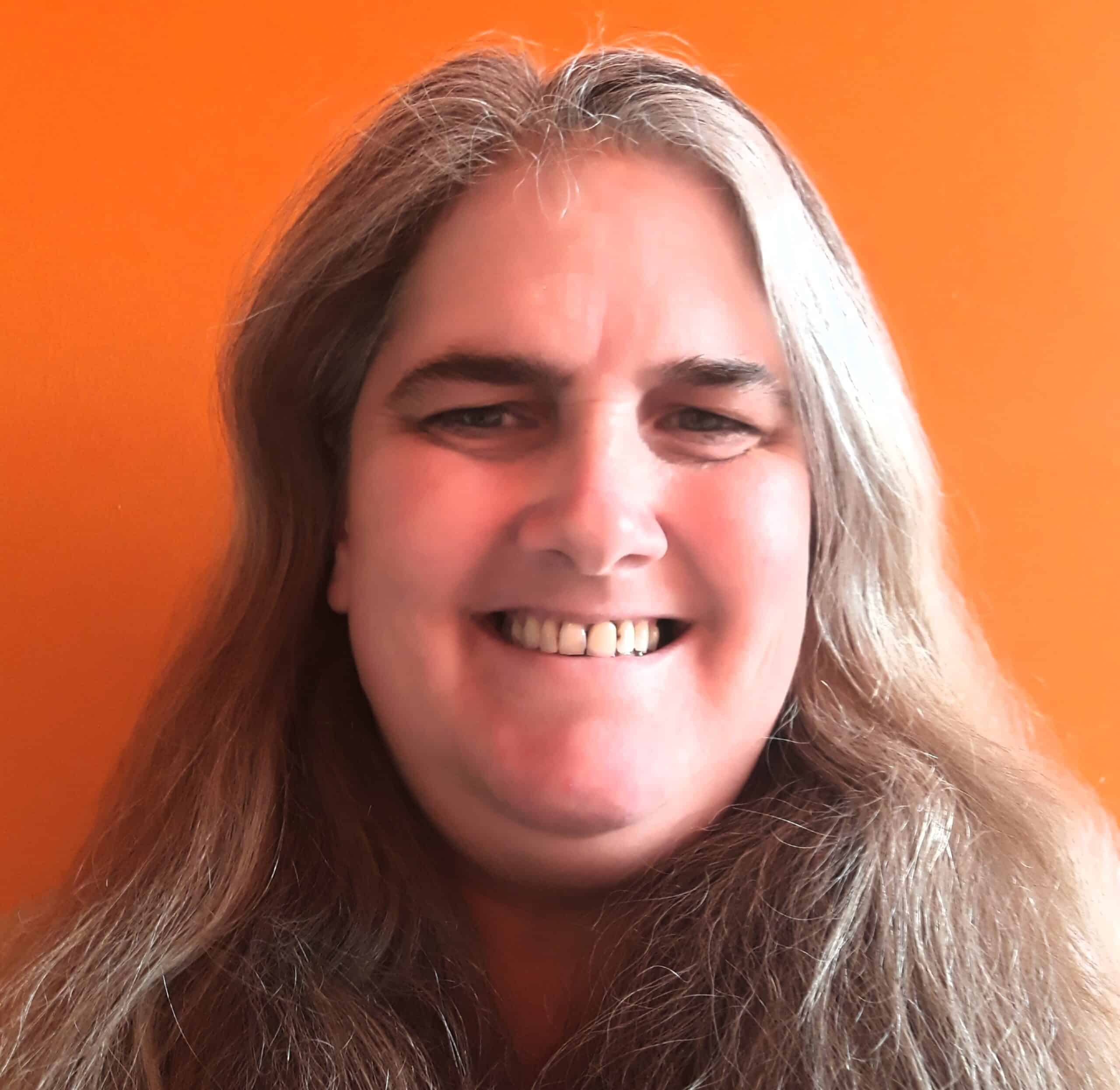 A person with long, wavy grey hair is smiling at the camera in front of a bright orange background, embodying the friendly face of National Curriculum education.