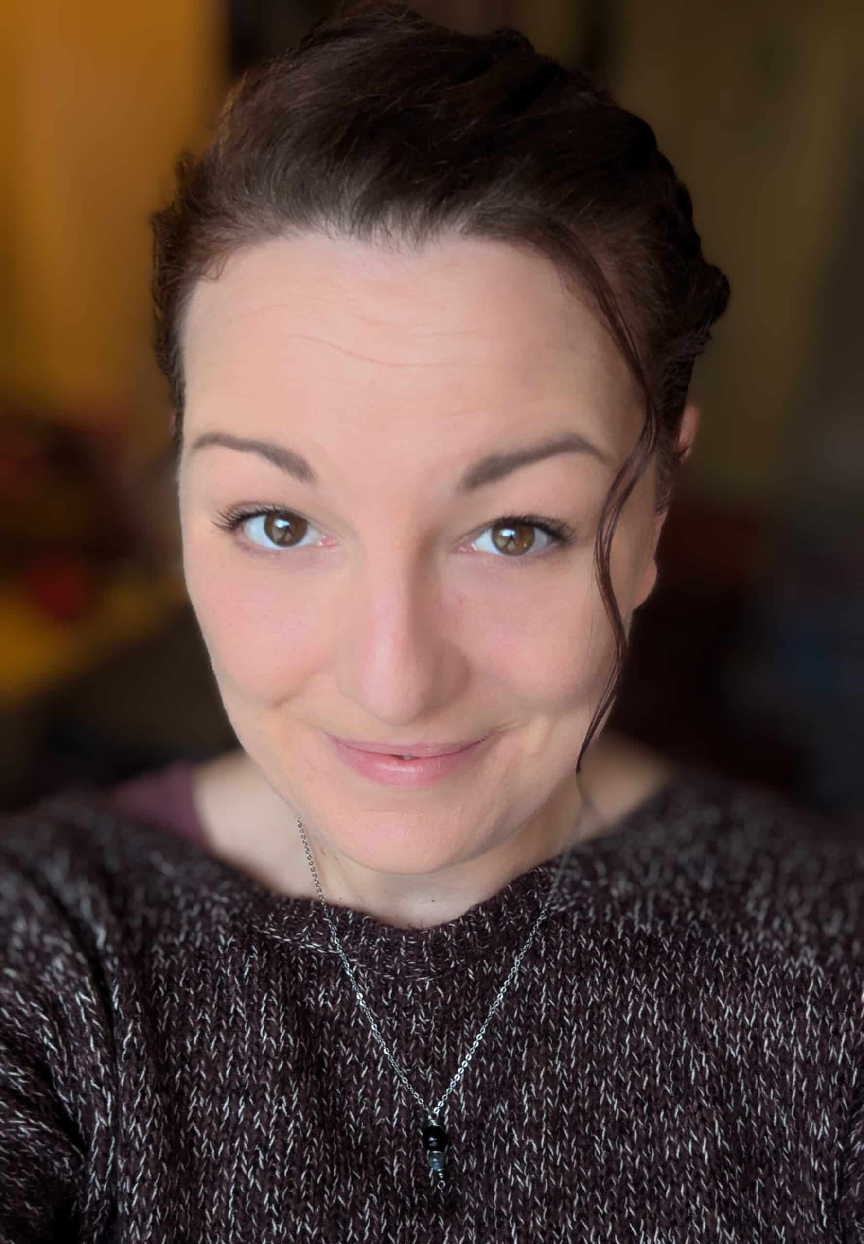 Kelly Pearson, a woman with fair skin and brown hair, wearing a dark knitted jumper and a pendant necklace, smiles softly at the camera. The background is softly blurred with warm tones.