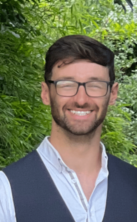 Patrick McAnea, a man with short dark hair, beard, and glasses, smiles outdoors in a light shirt and dark waistcoat, with green foliage in the background.