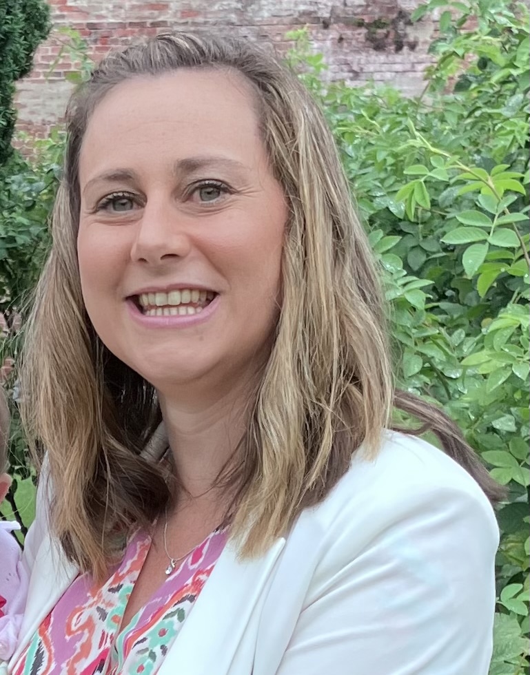 Emily McWatt, a smiling woman with shoulder-length light brown hair, stands outdoors in front of green foliage and a brick wall, wearing a white blazer and a colourful patterned top.