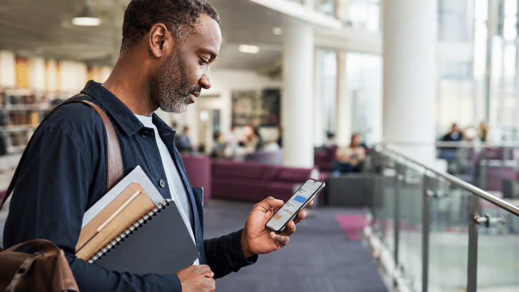 A man stands in a modern building, holding notebooks and a mobile, looking at the screen—perhaps reviewing some educational writing 📚. The background shows large windows, glass railings, and several people sitting on purple sofas, creating a suitable environment for study or collaboration.
